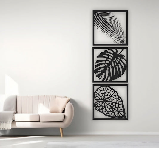 Three-piece vertical black wooden wall art set with palm, monstera, and vine leaf designs, mounted beside a cream sofa on a white wall.