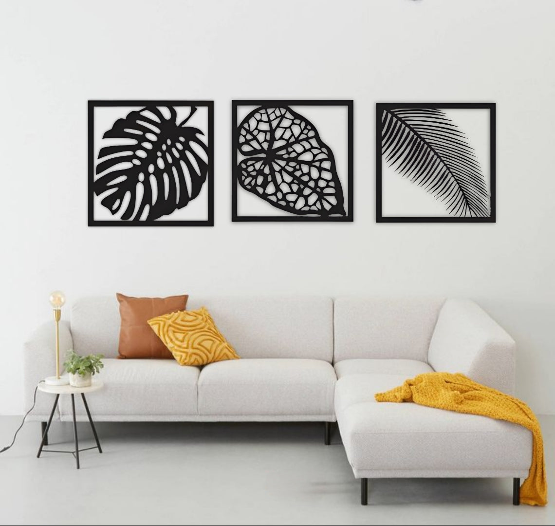 Three-piece vertical black wooden wall art set with palm, monstera, and vine leaf designs, mounted beside a cream sofa on a white wall.
