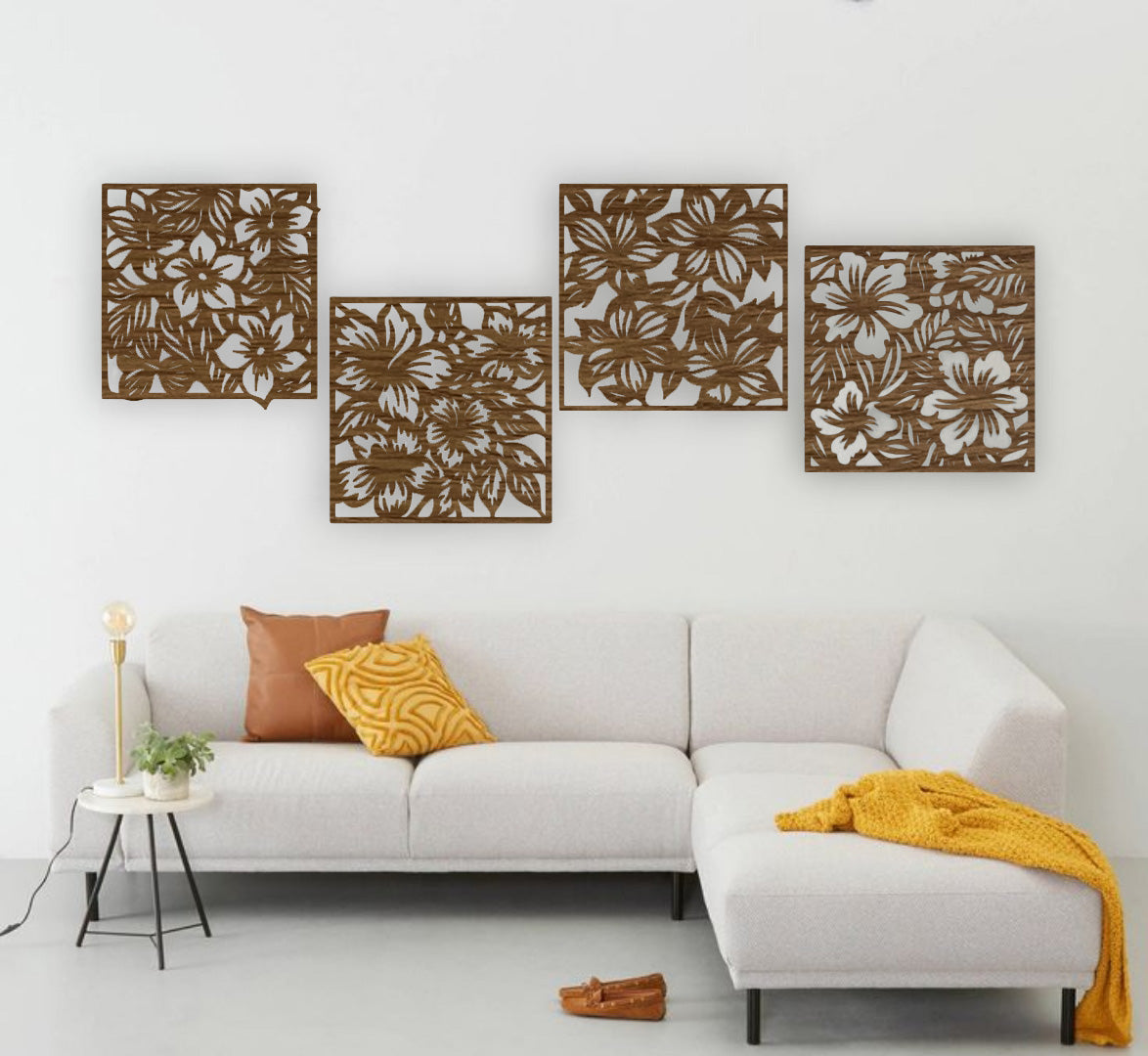 "Modern living room with a white L-shaped sofa and yellow accents, featuring a set of four black wooden laser cut floral wall art panels mounted on a white wall. Each panel displays intricate flower and leaf patterns, arranged in a staggered layout above the couch."