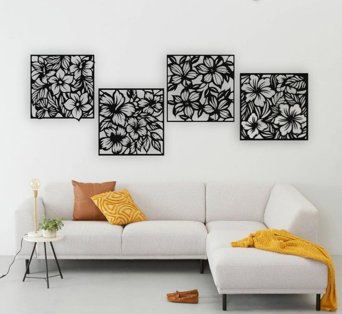 "Modern living room with a white L-shaped sofa and yellow accents, featuring a set of four black wooden laser cut floral wall art panels mounted on a white wall. Each panel displays intricate flower and leaf patterns, arranged in a staggered layout above the couch."