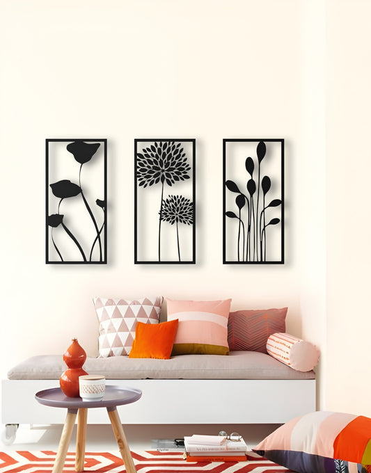 Three-piece botanical wooden wall art set in matte black finish, featuring floral silhouettes framed on a cream wall above a modern seating area with colorful pillows.