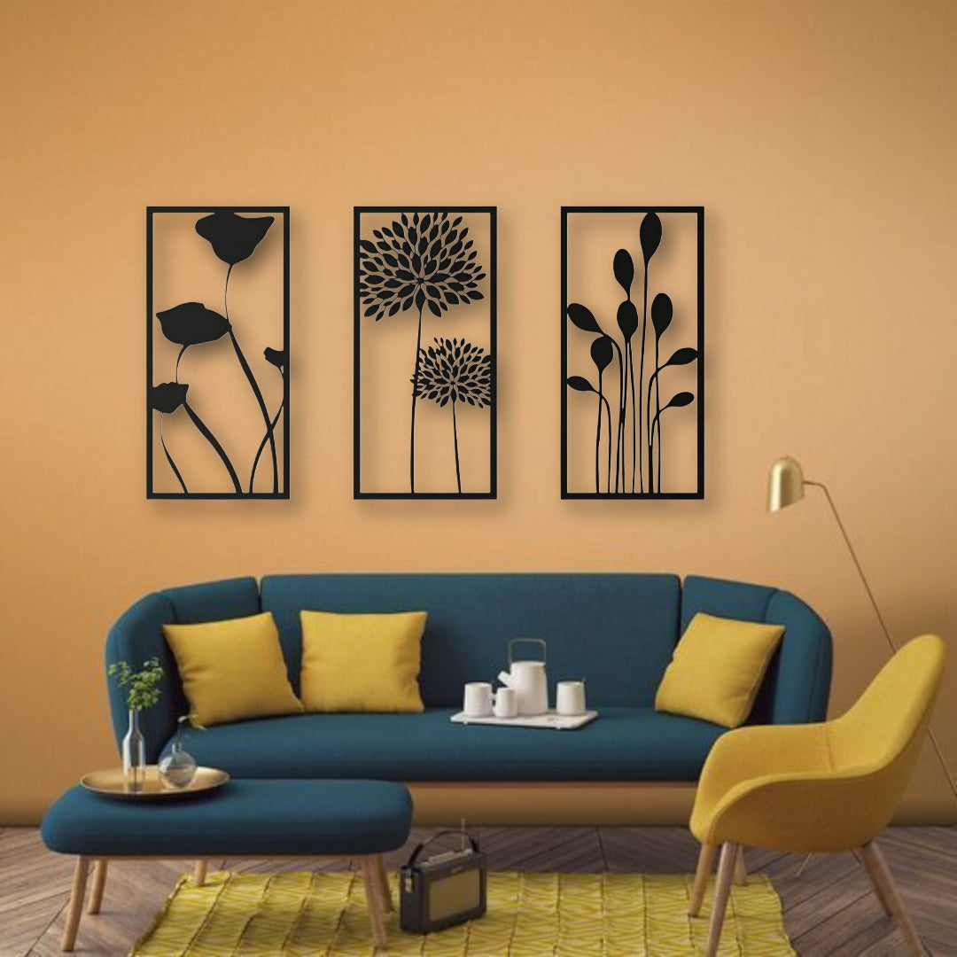 Three-piece botanical wooden wall art set in matte black finish, featuring floral silhouettes framed on a cream wall above a modern seating area with colorful pillows.