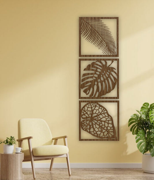 Three-piece vertical black wooden wall art set with palm, monstera, and vine leaf designs, mounted beside a cream sofa on a white wall.