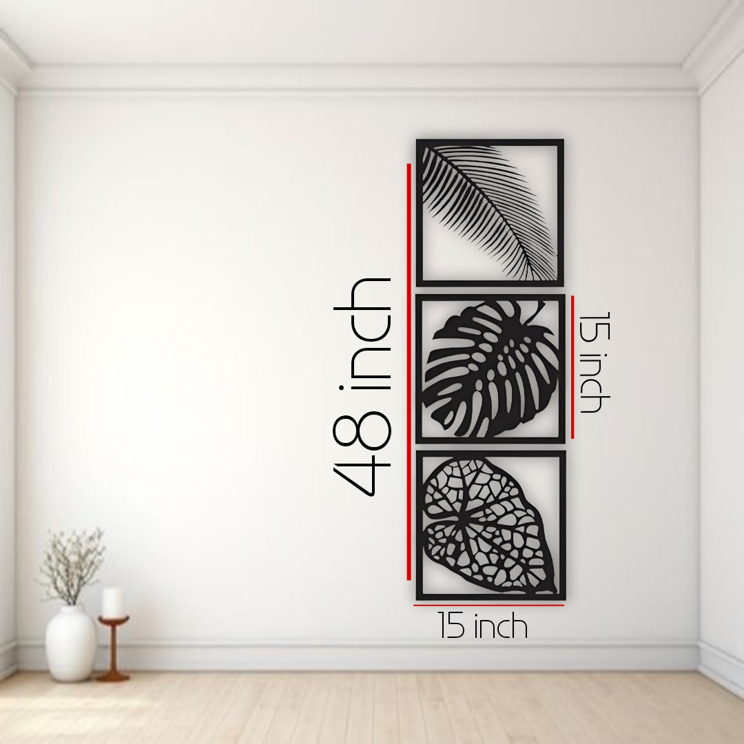 Three-piece vertical black wooden wall art set with palm, monstera, and vine leaf designs, mounted beside a cream sofa on a white wall.