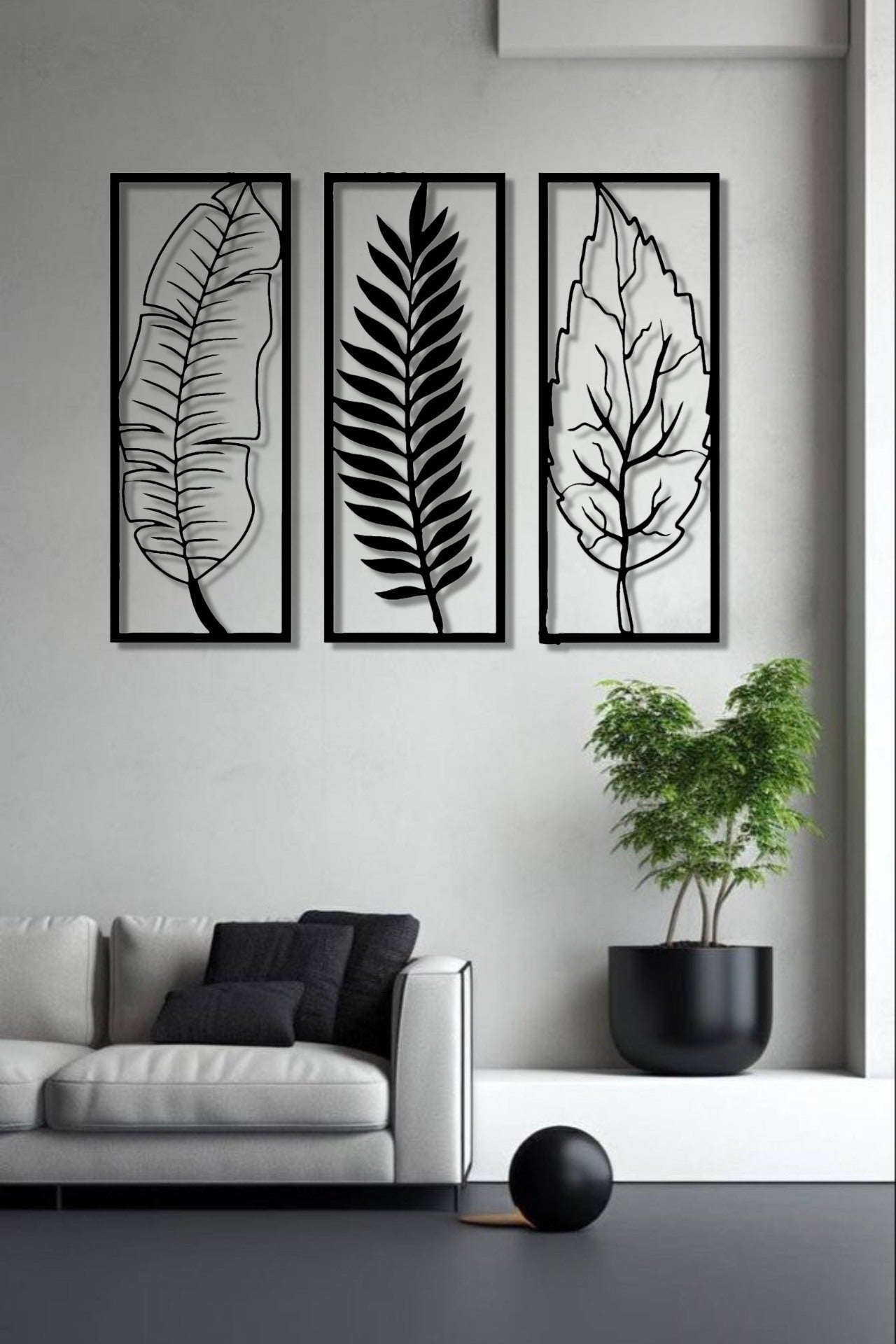 Set of three vertical wooden wall art panels in black finish featuring tropical leaves, mounted above a modern grey sofa in a minimalist living room